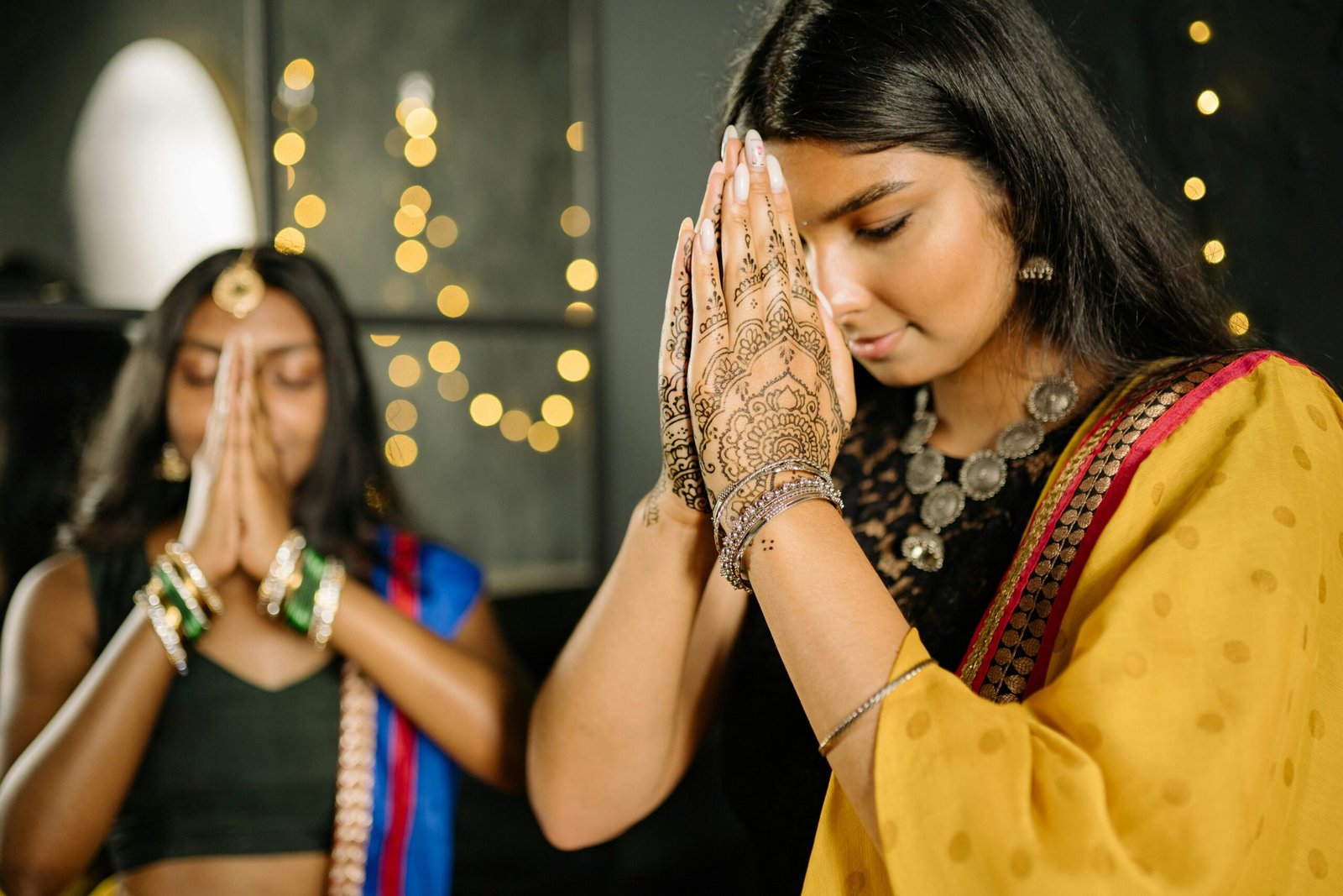 South Asian women in traditional attire and henna praying, celebrating Diwali indoors.
